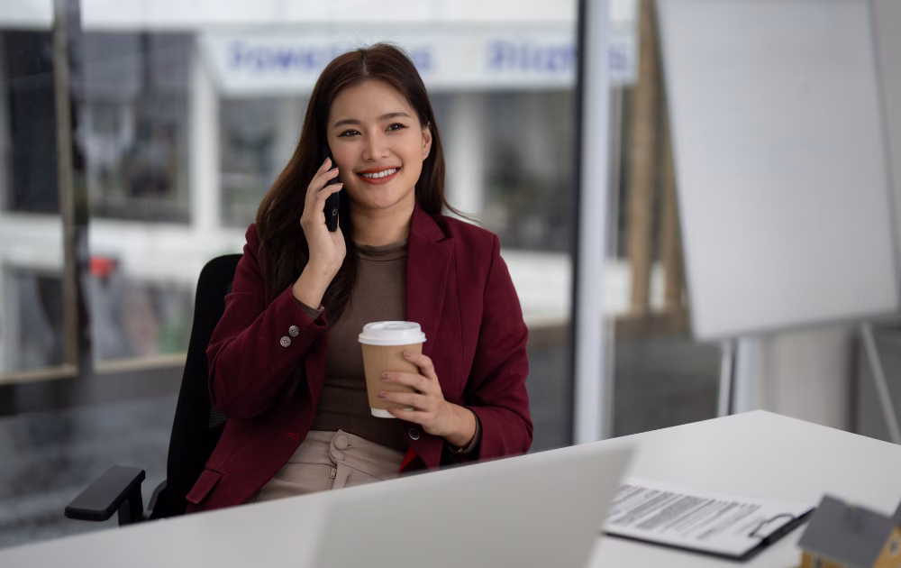 an asian woman in the office answering a phone call while holding a cup of coffee